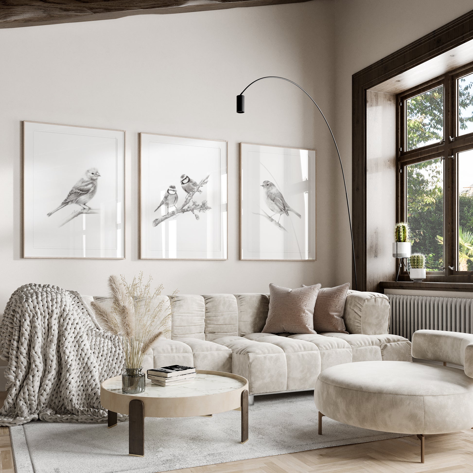 Set of 3 grey bird drawing art prints shown in frames hanging on a wall above a sofa. The British birds include a Robin Redbreast, Chaffinch and a pair of Blue Tit birds sitting on a branch. Art by Nicky Quartermaine Scott for Studio Q.