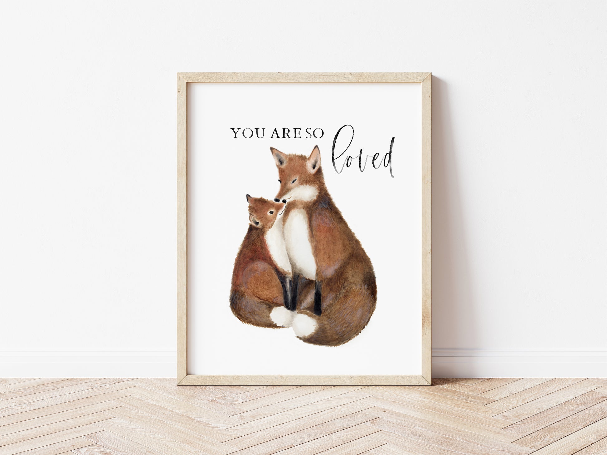 Painting of a mother and baby fox in rich rust, copper, browns and dark orange tones on a plain white background. Words You are So Loved are above the foxes. The picture is framed in a light wood frame and sits on a wood floor. Studio Q - Art by Nicky Quartermaine Scott
